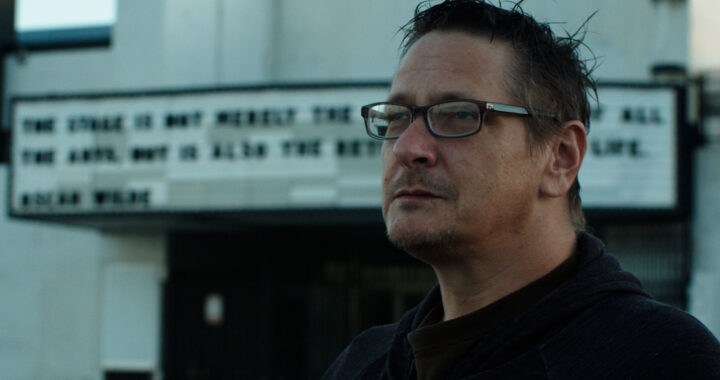 A white man in glasses stands in front of an old theater marquee.