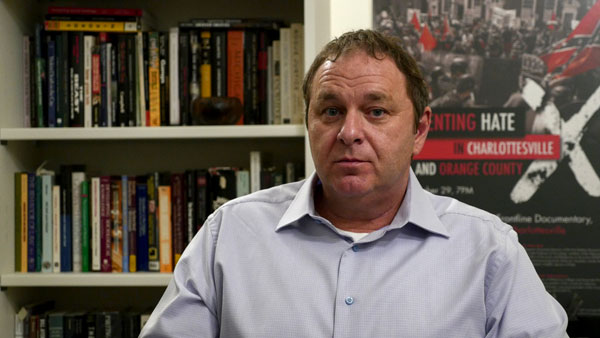 A white man in a blue button-down shirt sits pensively in front of a bookcase.